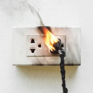 Picture of an electrical cord burst into flame while plugged into an electrical outlet.